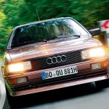 Klasyczny, brązowy Audi quattro z lat 80. pędzi po krętej, leśnej drodze.