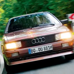 Klasyczny, brązowy Audi quattro z lat 80. pędzi po krętej, leśnej drodze.
