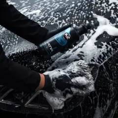 Ręce w czarnych rękawiczkach myją czarne auto pianą, trzymając butelkę szamponu.