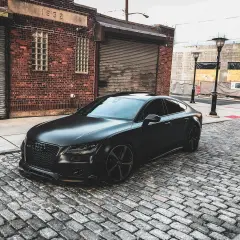 Matowy czarny Audi RS7 na brukowanej ulicy przed ceglanym budynkiem.