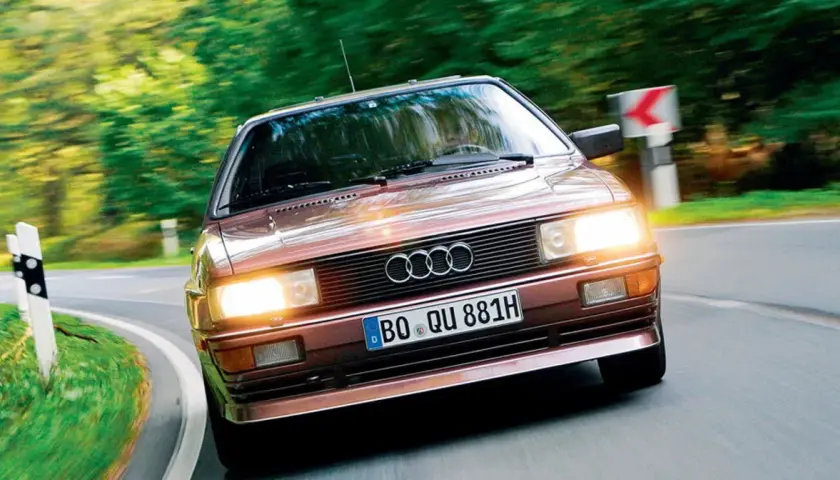 Klasyczny, brązowy Audi quattro z lat 80. pędzi po krętej, leśnej drodze.