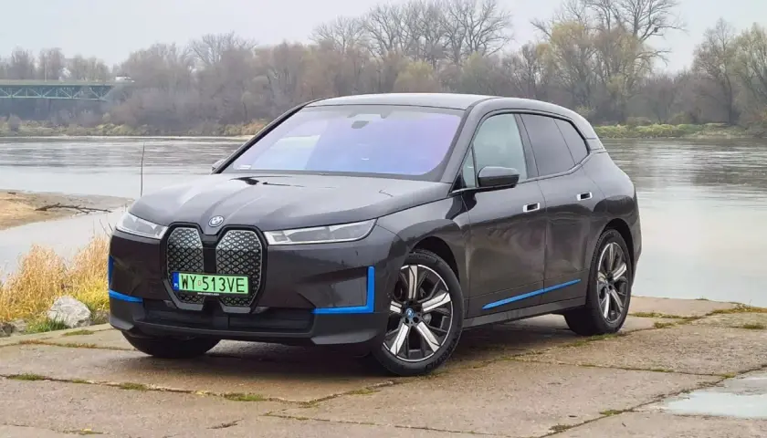 Ciemnoszary BMW iX z niebieskimi akcentami stoi nad rzeką, z mostem w tle.