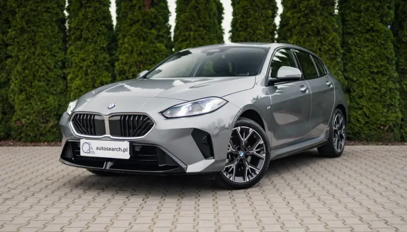 Szare BMW 118d z czarnym grillem i felgami stoi przed żywopłotem.
